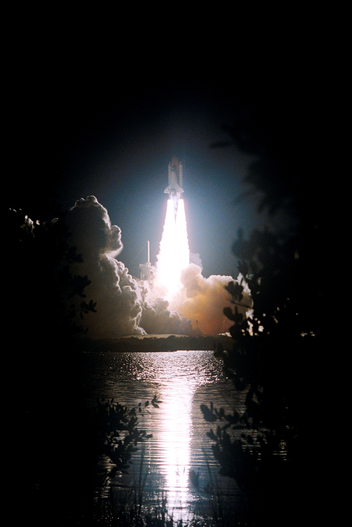 The fiery liftoff of Space Shuttle Discovery from Launch Pad 39B on mission STS-116 is reflected in the nearby water.